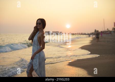 Eine ruhige Strandszene, in der eine Frau lächelt, während sie bei Sonnenuntergang entlang der Küste spaziert, mit fließenden Kleidern, sanften Wellen und einem Pier im Hintergrund, der die friedliche Atmosphäre am Meer unterstreicht. Stockfoto