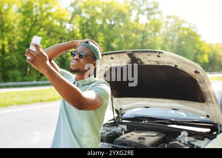 Verärgert schwarzen Kerl in der Nähe gebrochenen Auto mit offener Haube, Probleme, versucht, mobiles Netzwerk zu finden, ohne Signal Stockfoto