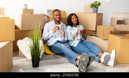 Glückliche afroamerikanische Ehegatten sitzen zwischen ausgepackten Kartons und trinken Kaffee und lächeln vor der Kamera Stockfoto