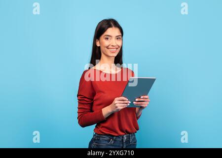 Frau in rotem Pullover lächelt, während sie die Tablette vor blauem Hintergrund hält Stockfoto