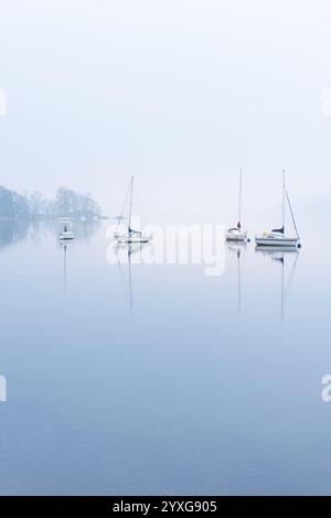 Ruhiger See mit spiegelartigen Reflexen und Yachten an einem nebeligen Morgen. Windermere, Lake District, Großbritannien. Stockfoto