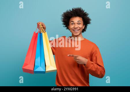 Der junge Mann zeigt aufgeregt bunte Einkaufstaschen vor blauem Hintergrund Stockfoto