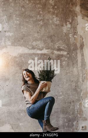 Dynamische Frau vor einer grauen Betonmauer mit einem kleinen Weihnachtsbaum in der Hand Stockfoto