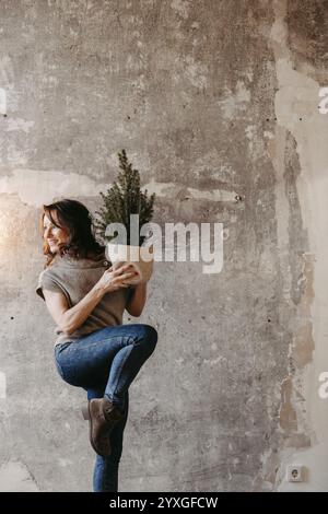 Dynamische Frau vor einer grauen Betonmauer mit einem kleinen Weihnachtsbaum in der Hand Stockfoto