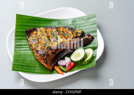 Ikan Pari Bakar oder gegrillter Sting Ray in Chili-Currysauce serviert auf Bananenblatt mit Schalotten, Gurke und Chili. Stockfoto