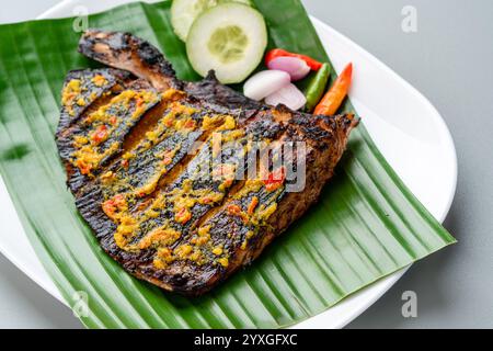 Ikan Pari Bakar oder gegrillter Sting Ray in Chili-Currysauce serviert auf Bananenblatt mit Schalotten, Gurke und Chili. Stockfoto