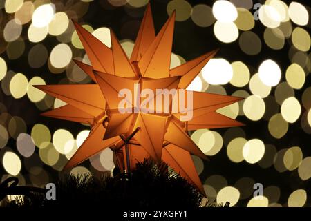 Ein orangener, weihnachtlicher Stern glänzt im Vordergrund vor einem wunderschönen, verschwommenen Bokeh-Hintergrund mit viel glitzerndem, funkelndem Licht Stockfoto