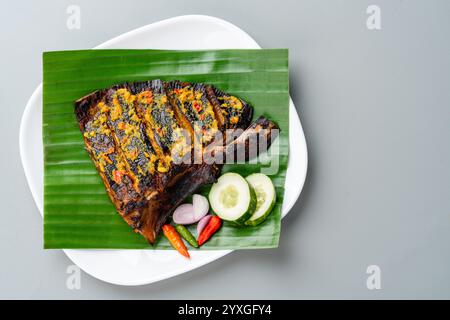 Ikan Pari Bakar oder gegrillter Sting Ray in Chili-Currysauce serviert auf Bananenblatt mit Schalotten, Gurke und Chili. Stockfoto