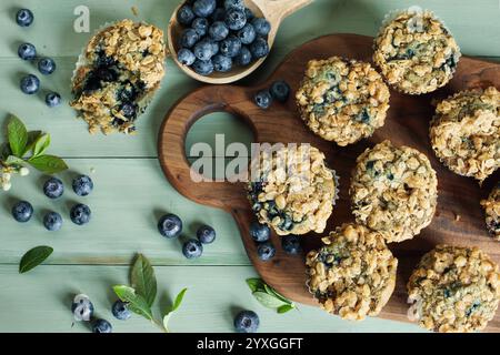 Frische Blaubeeren-Muffins mit Haferstreusel-Belag und rohe Blaubeeren, die von einem Holzlöffel verschüttet werden. Tischansicht mit Fokus auf Muffinplatten. Stockfoto