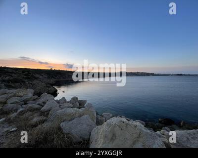 Dieses ruhige Küstenfoto fängt eine friedliche Szene während des Sonnenuntergangs in Zypern ein. Das ruhige Meer reflektiert die warmen Farbtöne der verblassenden Sonne, während der Teppich Stockfoto