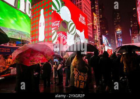 Besucher des Times Square in New York am Mittwoch, den 11. Dezember 2024, spazieren im Regen unter den digitalen Plakaten mit Weihnachtsmotiven, die Coca-Cola Werbung machen. (© Richard B. Levine) Stockfoto