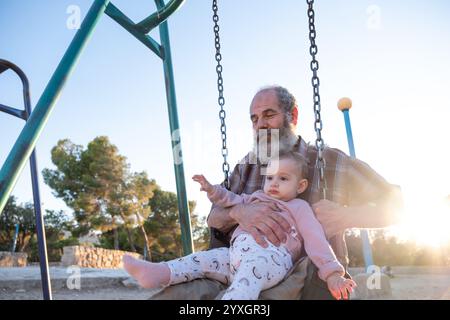 Ein älterer Mann teilt einen freudigen Moment mit einem Baby auf einer Schaukel im Park während Sonnenuntergang. Die warme Beleuchtung und der malerische Hintergrund halten das Wesen von fest Stockfoto