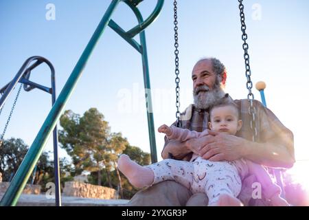 Ein älterer Mann teilt einen freudigen Moment mit einem Baby auf einer Schaukel im Park während Sonnenuntergang. Die warme Beleuchtung und der malerische Hintergrund halten das Wesen von fest Stockfoto