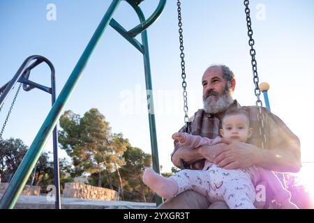 Ein älterer Mann teilt einen freudigen Moment mit einem Baby auf einer Schaukel im Park während Sonnenuntergang. Die warme Beleuchtung und der malerische Hintergrund halten das Wesen von fest Stockfoto