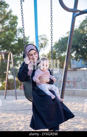 Ein herzerwärmender Moment, in dem eine muslimische Mutter im traditionellen Abaya- und Hijab-Stil gefangen wird, die Freude am Spielplatz mit ihrem Baby teilt und kulturelle vielfalt miteinander verbindet Stockfoto