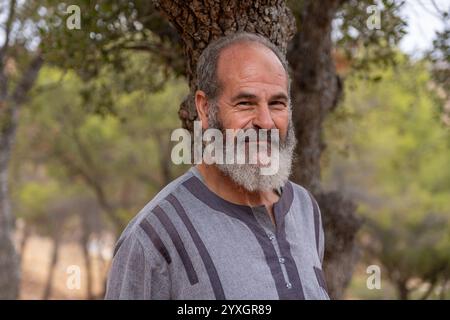 Ein älterer Mann mit einem warmen Lächeln, der an einem Baum in einem ruhigen Wald steht. Sein friedlicher Ausdruck und die natürliche Umgebung vermitteln ein Gefühl der Ruhe Stockfoto