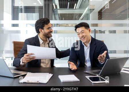 Zwei fröhliche Profis sitzen an einem modernen Bürotisch und führen ein lebhaftes Gespräch. Die Umgebung deutet auf eine positive Zusammenarbeit hin, mit Dokumenten und Laptops, die auf Arbeitsaktivitäten hinweisen Stockfoto