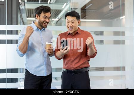 Zwei Büroangestellte feiern einen Erfolg in der Nähe eines modernen Bürofensters. Sie sind begeistert und teilen einen freudigen Moment, wenn sie etwas Positives auf einem Smartphone ansehen. Stockfoto