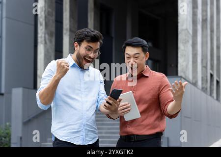 Zwei fröhliche Kollegen reagieren fröhlich auf gute Nachrichten mit einem Smartphone im Freien. Ihre jubelnden Ausdrucksformen vermitteln Teamarbeit, Leistung und Glück in einem professionellen Umfeld Stockfoto