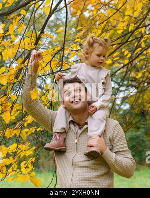 Fröhlicher Vater und kleine Tochter spielen während eines Spaziergangs durch den wunderschönen Herbstpark. Mädchen, das auf Daddys Schultern sitzt. Perfekter Wochenendvater mit seinem du Stockfoto