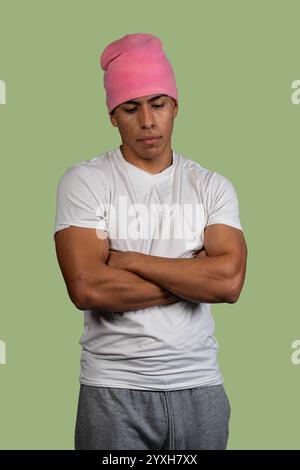 Studio-Porträt eines ernsthaften jungen latinos mit rosa Beanie, weißem T-Shirt und grauer Jogginghose, die Arme kreuzt und nach unten blickt Stockfoto