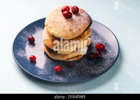 Hüttenkäsepfannkuchen mit Preiselbeeren werden auf einer blauen Platte auf einem hellblauen Tisch gestapelt, mit Puderzucker bestreut. Hochwertige Fotos Stockfoto