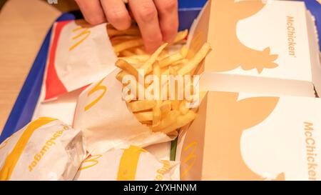 Hand aufsammeln Pommes Frites vom Fast-Food-Tablett mit McDonald's Burgern und McHühnerboxen Nahaufnahme Detail Casual Dining Konzept, ungesundes Essen, l Stockfoto