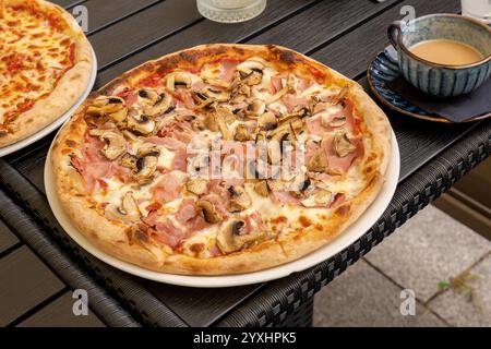 Köstliche, frische Pilz- und Schinkenpizza, serviert auf einem Restauranttisch im Freien neben einer Keramikkaffee, Nahaufnahme von italienischen Cuis Stockfoto