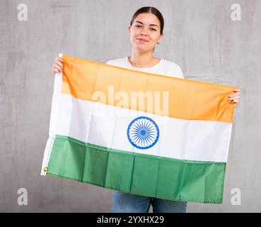 Fröhlich lächelnde junge Frau, die Flagge Indiens vor grauem Hintergrund schwenkt Stockfoto