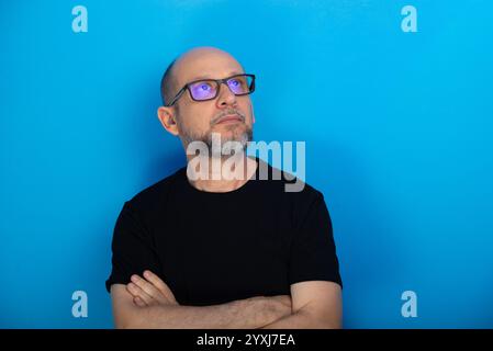 Bärtiger, kahler Mann mit verschreibungspflichtiger Brille und überkreuzten Armen posiert zum Foto vor blauem Hintergrund. Studio-Aufnahme. Stockfoto