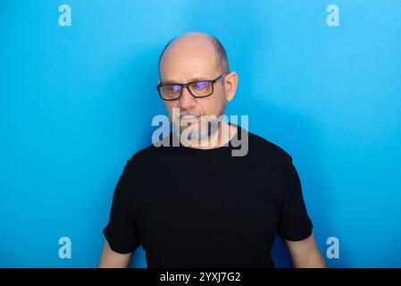 Bärtiger, kahler Mann, der eine Brille trägt und ein schwarzes Hemd, das vor blauem Hintergrund für ein Foto posiert. Studio-Aufnahme. Stockfoto