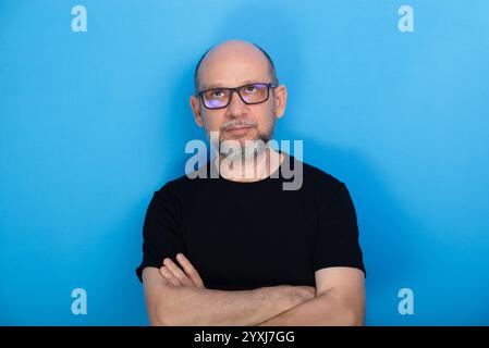 Bärtiger, kahler Mann mit verschreibungspflichtiger Brille und überkreuzten Armen posiert zum Foto vor blauem Hintergrund. Studio-Aufnahme. Stockfoto
