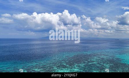 Eine atemberaubende tropische Meereslandschaft mit kristallklarem azurblauem Wasser, einer fernen üppigen Insel und dramatischen weißen Wolken vor einem leuchtend blauen Himmel. Pe Stockfoto