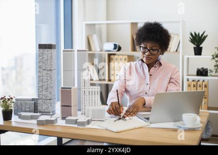 Afrikanische Architektin im Büro, die Wolkenkratzermodelle für moderne Stadt entwirft. Junger Profi, der Architekturpläne erstellt, während er an einem Notebook arbeitet. Stockfoto