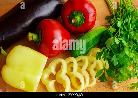 Buntes Gemüse, einschließlich Auberginen, Paprika, Gurke und Koriander, wird auf einem hölzernen Schneidebrett ordentlich angeordnet, bereit zum Zerkleinern und Kochen Stockfoto