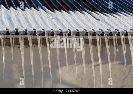 Scharfe Eiszapfen und geschmolzener Schnee hängen von den Dachtrassen. Wunderschöne, transparente Eiszapfen, die langsam über ein Dach gleiten. Stockfoto