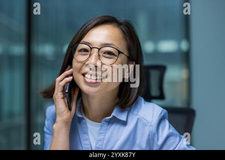 Asiatische Frau, die während ihrer Arbeit in einem Büro telefonierte. Sie zeigt eine positive Einstellung und stellt ein geschäftsorientiertes, aber ansprechbares Verhalten dar. Stockfoto