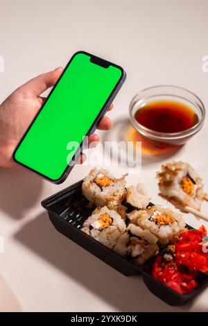 Frau, die zu Hause Sushi-Brötchen isst. Smartphone mit grünem Bildschirm. Kopierraum für Restaurants, Lebensmittellieferungen, Food-Websites und A Stockfoto