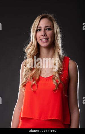 Eine blonde Frau in einem orangefarbenen Kleid, gefangen in einem halblangen Porträt. Sanftes Lächeln, entspannte Arme, schwarzer Hintergrund, mit Licht von rechts akzentuiert Stockfoto
