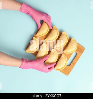 Flach liegen die Hände einer Bäckerin in rosa Gummihandschuhen legt rohe Kuchen auf blauem Hintergrund, gefrorene Halbzeuge Stockfoto