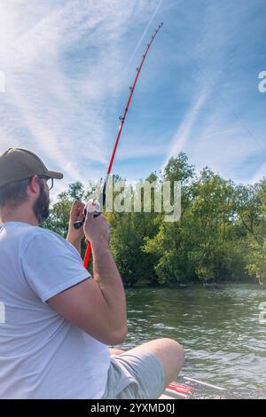 Ein Mann fischt mit einer roten Rute und einem weißen Hemd. Der Mann sitzt auf einem Boot im Wasser Stockfoto