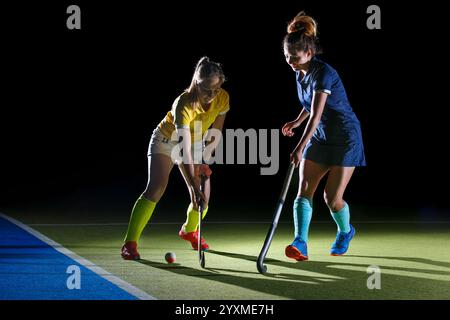 Zwei weibliche Feldhockeyspielerinnen, eine in einem gelben Hemd und die andere in Blau, treten nachts in einem Wettkampf mit leuchtender Feldbeleuchtung an Stockfoto