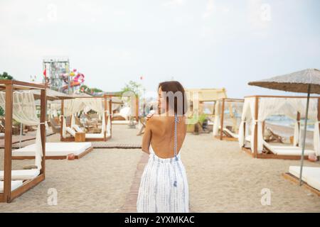 Eine fröhliche Frau in fließendem Sommerkleid spaziert auf einem Sandweg in einem luxuriösen Strandresort. Umgeben von eleganten hölzernen Cabanas, fängt die Szene eine ruhige und stilvolle Urlaubsatmosphäre ein. Stockfoto