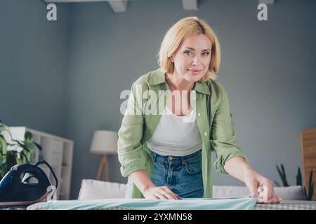 Reife Frau mit blonden Haaren, die Hausarbeit in einem gemütlichen Wohnzimmer macht und ein lässiges grünes Hemd trägt, während sie zu Hause Hausaufgaben erledigt Stockfoto