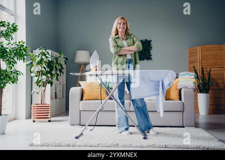 Selbstbewusste Frau, die im gemütlichen Wohnzimmer bügelt, lässiges Outfit trägt, Freude ausdrückt, während sie sich mit der Hausreinigung und der Hausarbeit beschäftigt. Stockfoto
