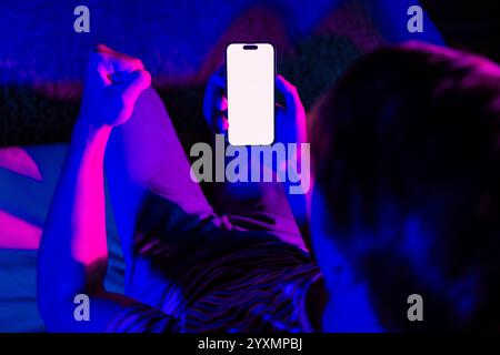 Junger Mann, der ein Smartphone mit leerem Bildschirm hält, beleuchtet von leuchtendem rosa und blauem Neonlicht in einer häuslichen Umgebung Stockfoto