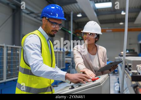 Die indische Betriebsleiterin trainiert einen afroamerikanischen männlichen Mitarbeiter in einem blauen Helm und einer gelben Weste, um Maschinen zu bedienen. Stockfoto