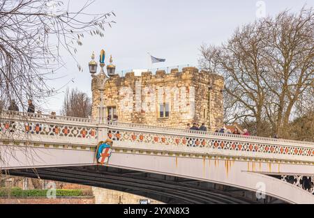 Eine Nahaufnahme einer historischen Eisenbrücke mit einem alten Turm im Hintergrund. Die Menschen sind auf der Brücke und Winterbäume auf beiden Seiten. Stockfoto