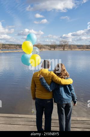 Unerkennbare Frau und Mann, umarmt einander, stehen mit dem Rücken am Ufer des Sees und halten blaue gelbe Ballons. Tag der Einheit zur Unterstützung Großbritanniens Stockfoto