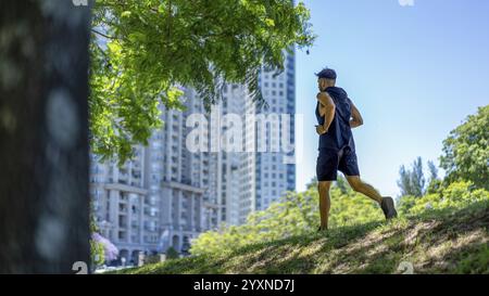 Junger, sportlicher Mann, der auf hügeligen Wegen in einem modernen Stadtpark, umgeben von Wolkenkratzern, läuft Stockfoto